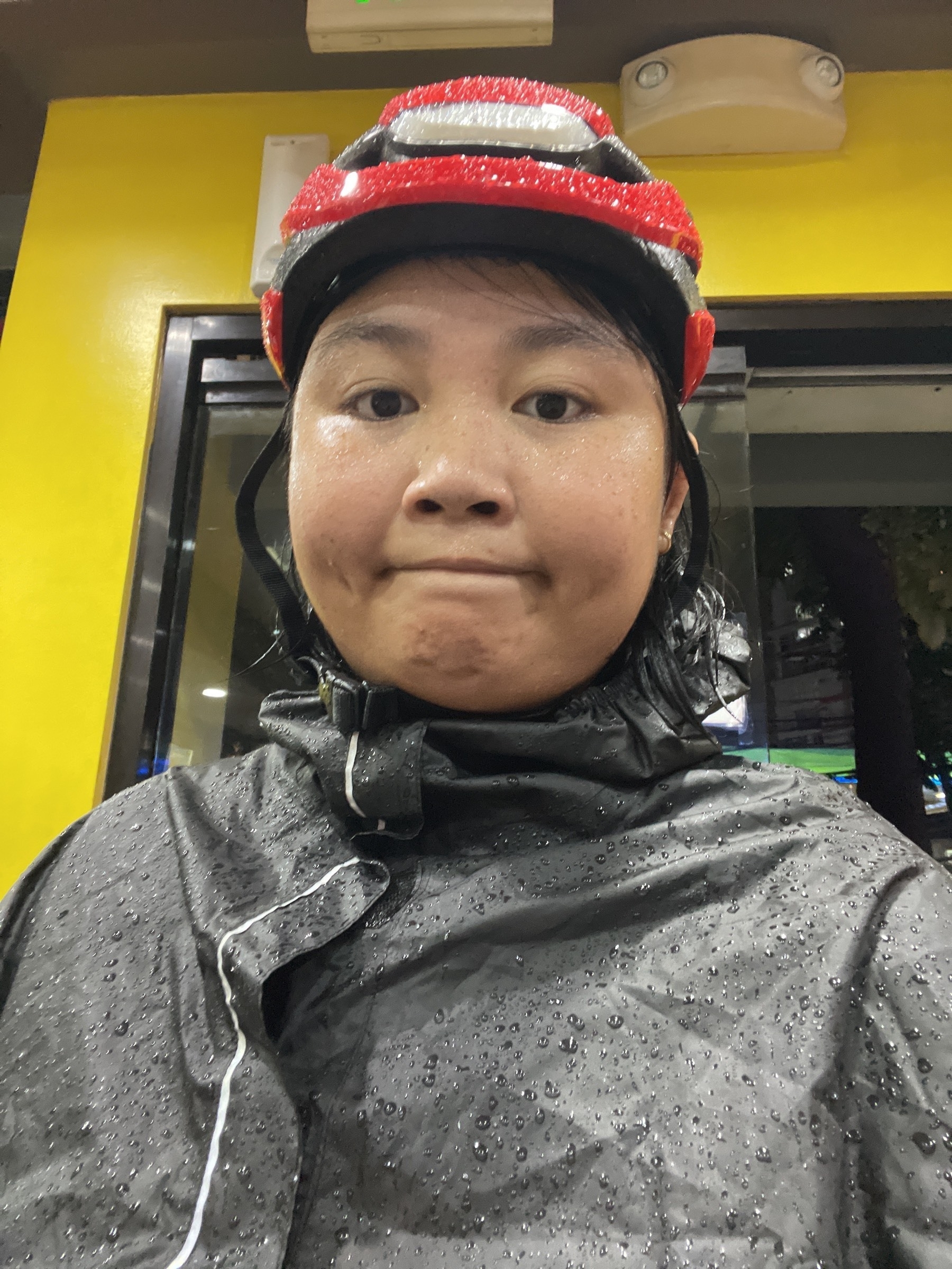 Selfie of Chi in her rain gear for her bike commute, wearing her red Lumos helmet and a black rain poncho. Both the poncho and her helmet are visibly drenched or have rain drops on them, and Chi's making a dejected or kind of exhausted face.