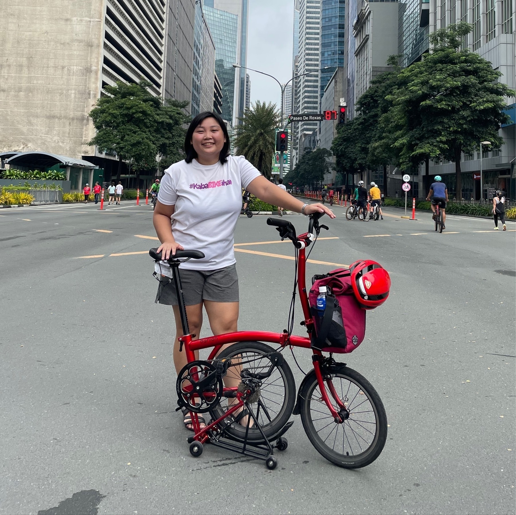 Chi smiling and posing with her red Banian trifold bike, which is partially folded, while in the middle of Ayala Avenue because it's a Car-Free Sunday morning event. She is wearing a white t-shirt that has the text, "#KabaBIKEihan" printed, and gray shorts and black sandals. On her bike, her red GoFar front mounted bag is attached, and her red Lumos helmet is also strapped to her bag.