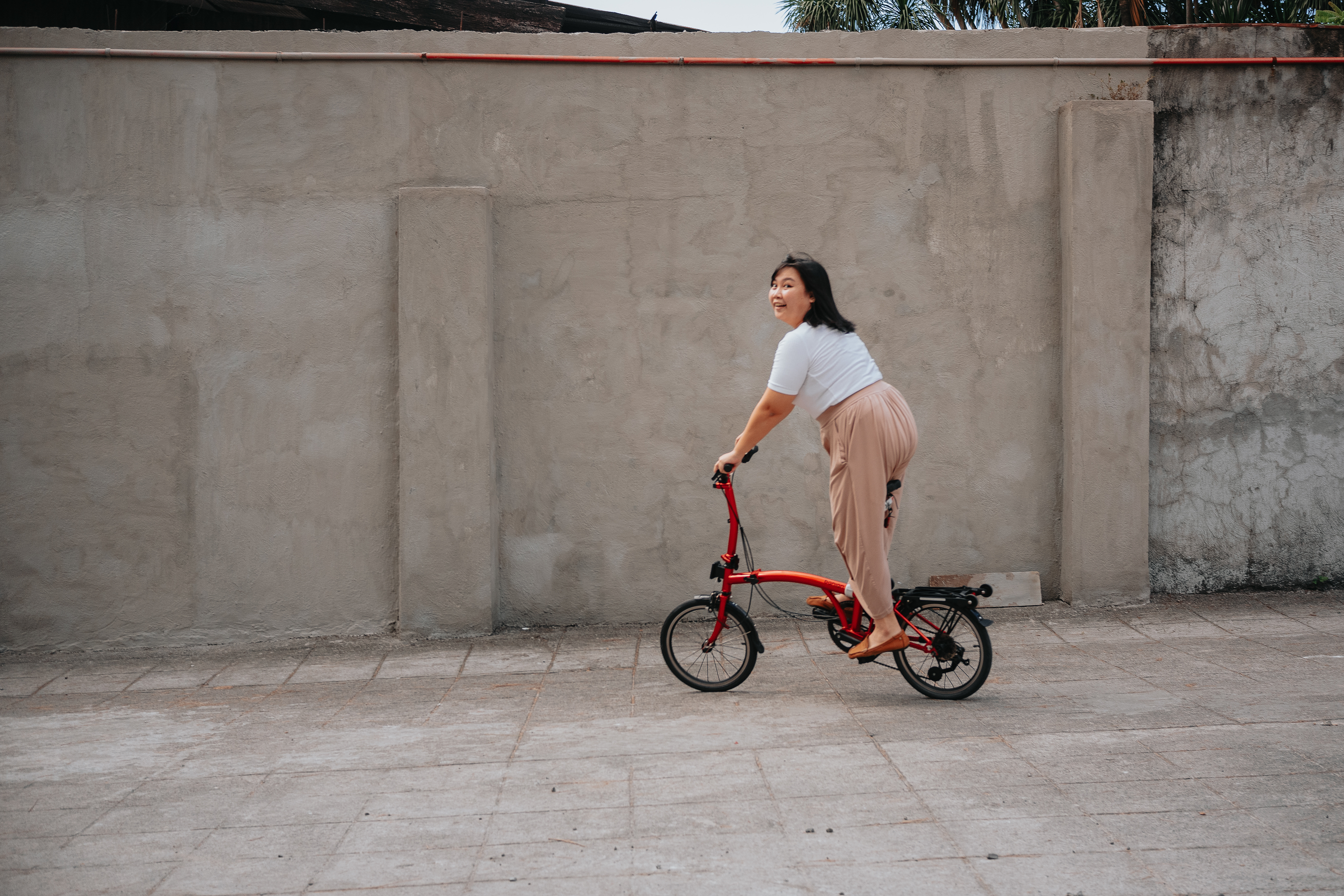Chi riding her red Banian trifold while wearing a white top, a long beige flowy skirt, and brown loafers. Her expression shows that she's having fun, while turning to look at the camera. She's not fully seated on the bike, and instead is coasting on it while turning to go towards the left.