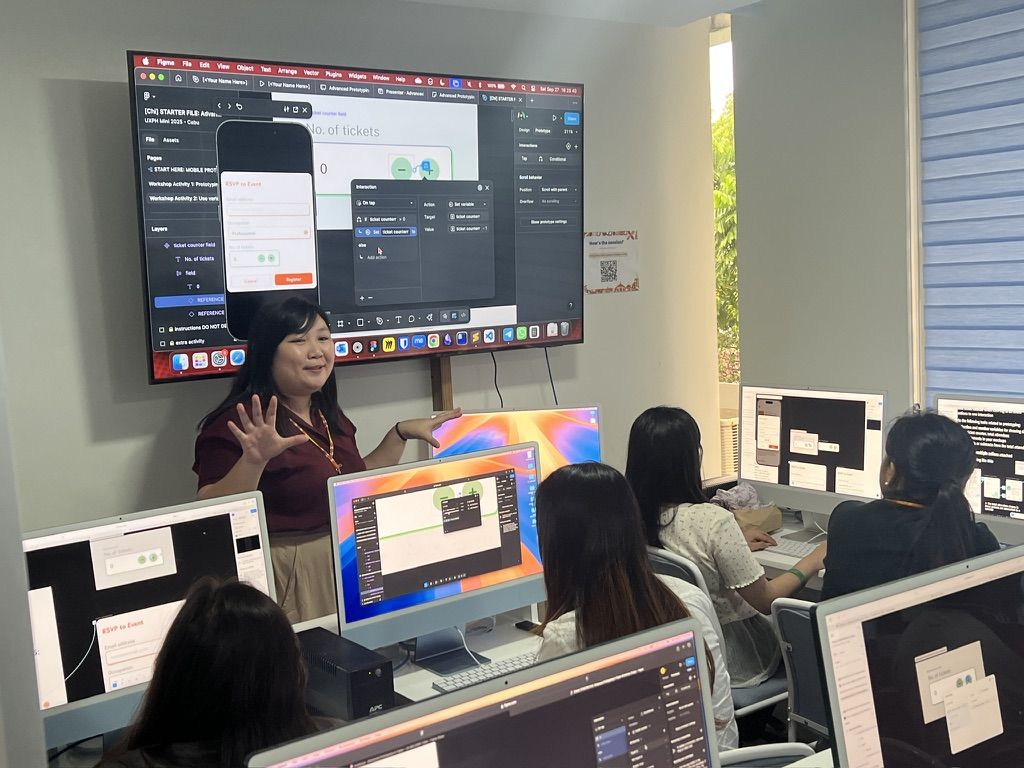 This classroom was super cramped and I did my best to be seen and heard with all my gesturing lolol Chi is standing in front of a small classroom, both her hands up as she explains what is shown on the big TV screen behind her, which is sharing her laptop’s screen with Figma open and the prototype preview overlay showing a modal within a phone frame. Attendees are positioned around and in front of her, with iMacs on each table and attendees also following along in Figma during the activity.