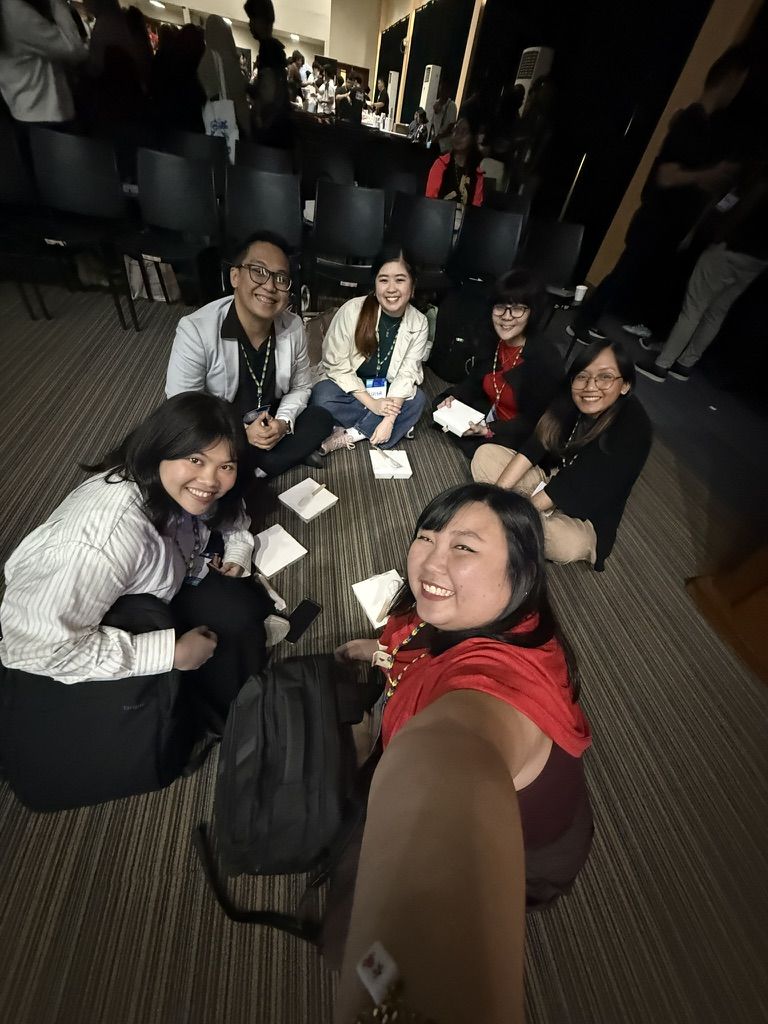 With speakers and workshop facilitators for UXPH Mini 2025: Davao! The first leg wooo 😁 Chi doing a wide angle selfie with other workshop facilitators and speakers from UXPH Mini 2025: Davao. They are seen sitting on the floor of an auditorium while eating lunch in a group huddle setup.