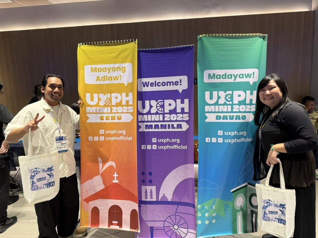 With Richard while at UXPH Mini 2025: Manila Chi and Richard posing in front of the 3 tarps for the different legs for UXPH Mini 2025. Richard is by the left side of the Manila leg tarp holding up a peace sign, and Chi is on the right side with the Davao leg tarp. Both Chi and Richard are also holding on to their conference swag bags.
