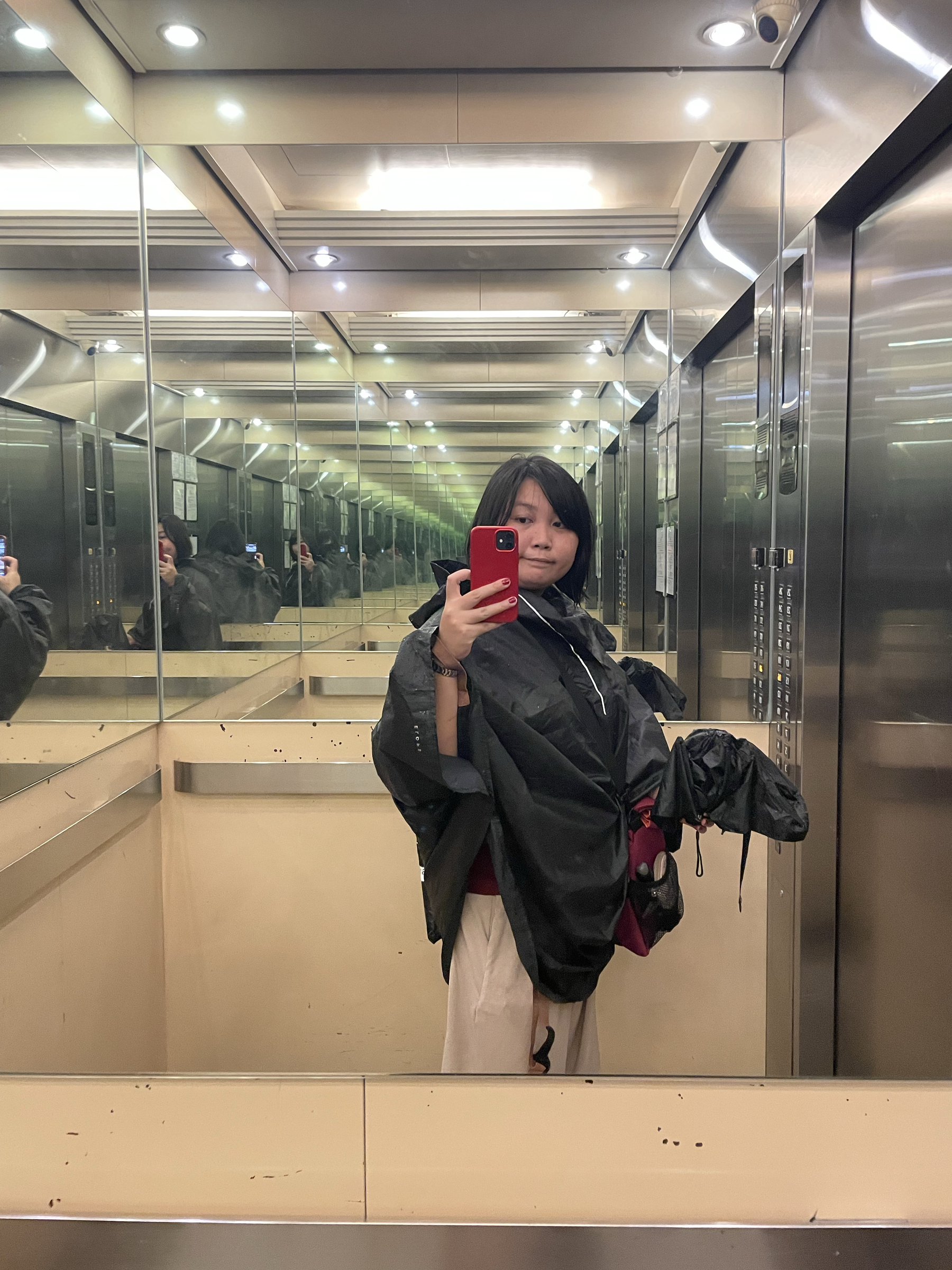 mirror photo of Chi in their condominium’s elevator. She is seen wearing her bike poncho over her work clothes. Her bike bag is in sling mode and set to her left, and in her left hand she is also seen holding a wet folded umbrella.