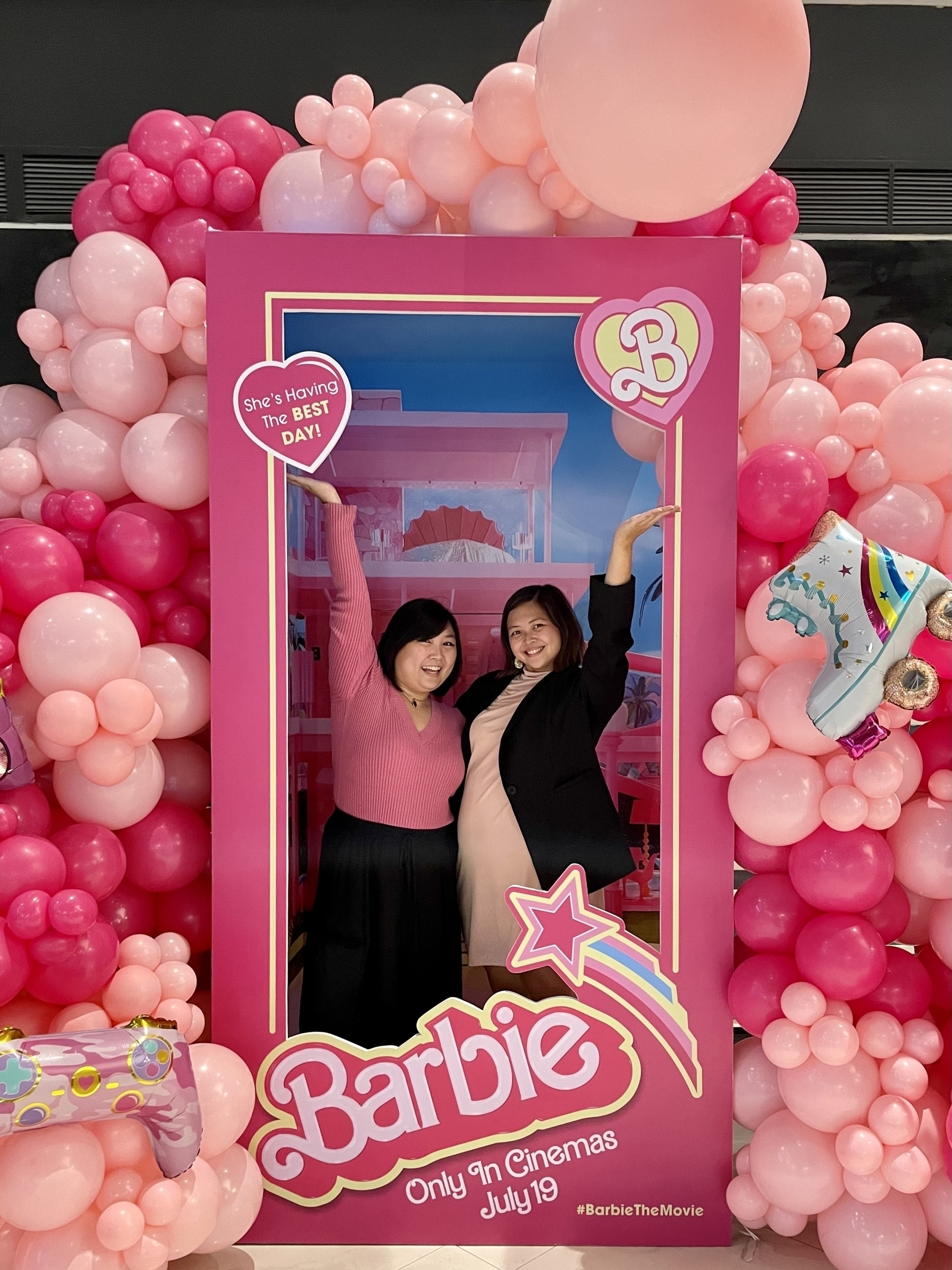 Chi and Karen posing in the Barbie Box promotional stand for the movie. Chi is dressed in a pink long-sleeved top and a black long skirt, while Karen is wearing a light pink turtleneck dress with a black blazer.