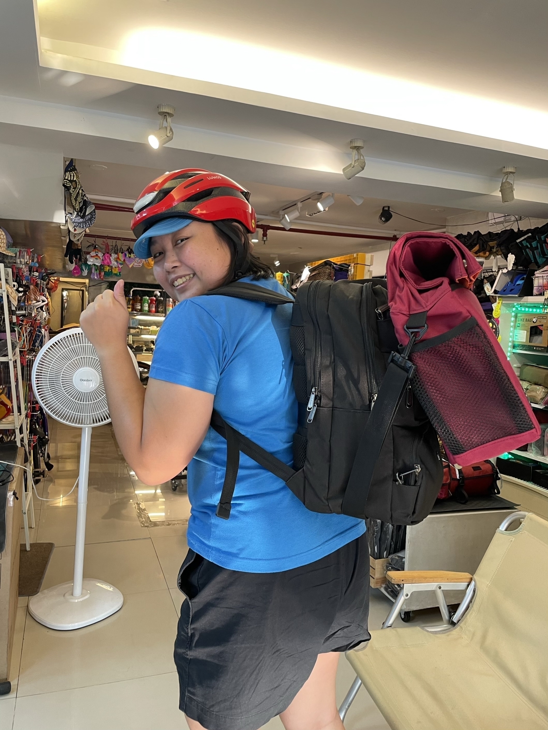 photo of Chi gesturing with a thumbs up to the camera with her backpack on, and her new front bag, meant for a trifold folding bike with a front carrier block, attached to the front of her backpack.