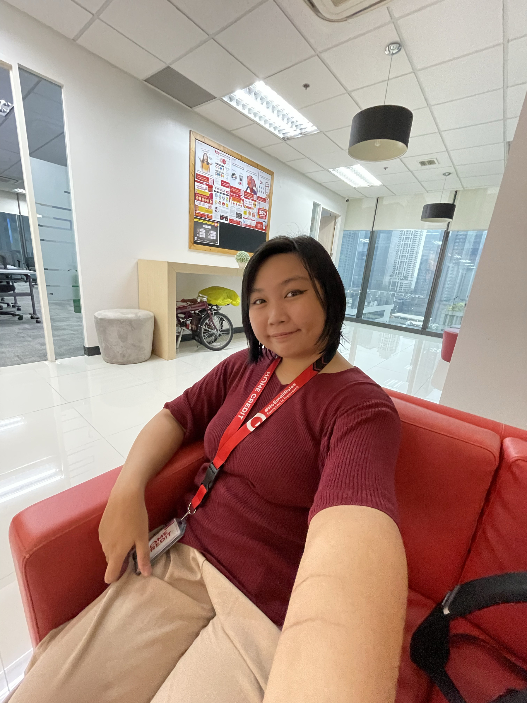 wide angle selfie of Chi in office attire sitting in their office lobby lounge, with her folding bike neatly tucked under a table and seen behind her.