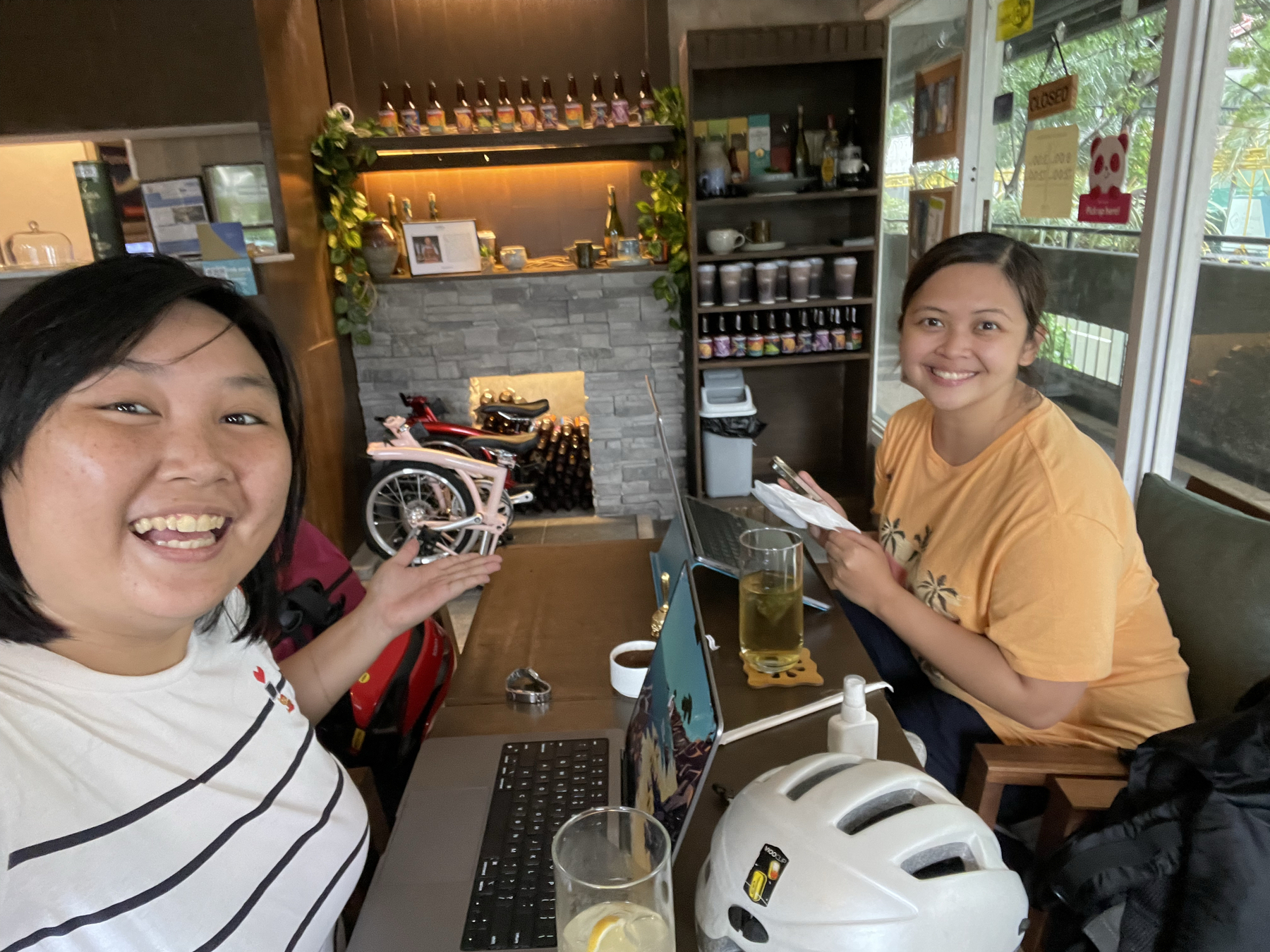Chi and Karen posing for a selfie. In the background you can see both Chi's red trifold and Karen's blush pink trifold folded in the background. On the table, both Chi and Karen have their laptops out, and are co-working.