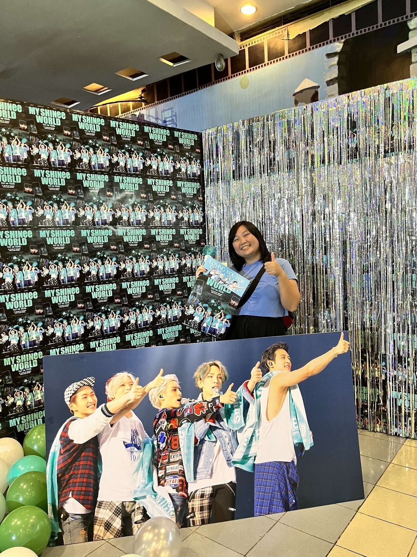 A photo of Chi with the MY SHINee WORLD movie poster backdrop and with freebies from the documentary premiere. In front of her are a photo of the SHINee members (from left to right): Onew, Taemin, Jonghyun, Key, and Minho all pointing a thumbs up to the right side during one of their concerts. Chi is also posing with a thumbs up to join the SHINee members.