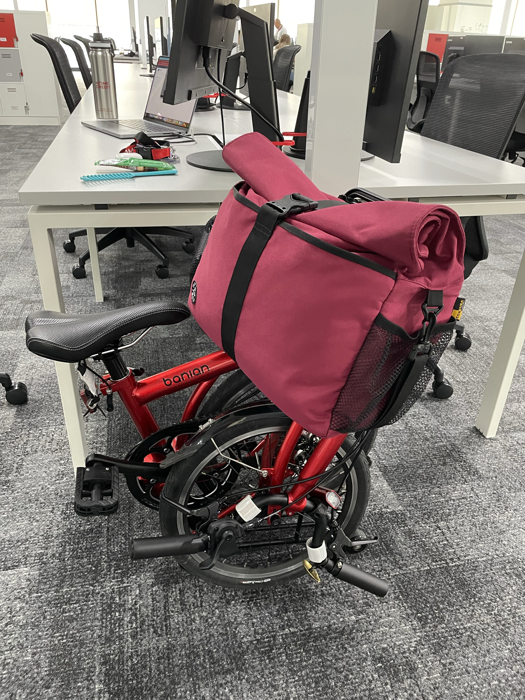 Chi’s trifold bike in the fully folded state positioned next to her table at the office. Her front bag is still attached to the bike.