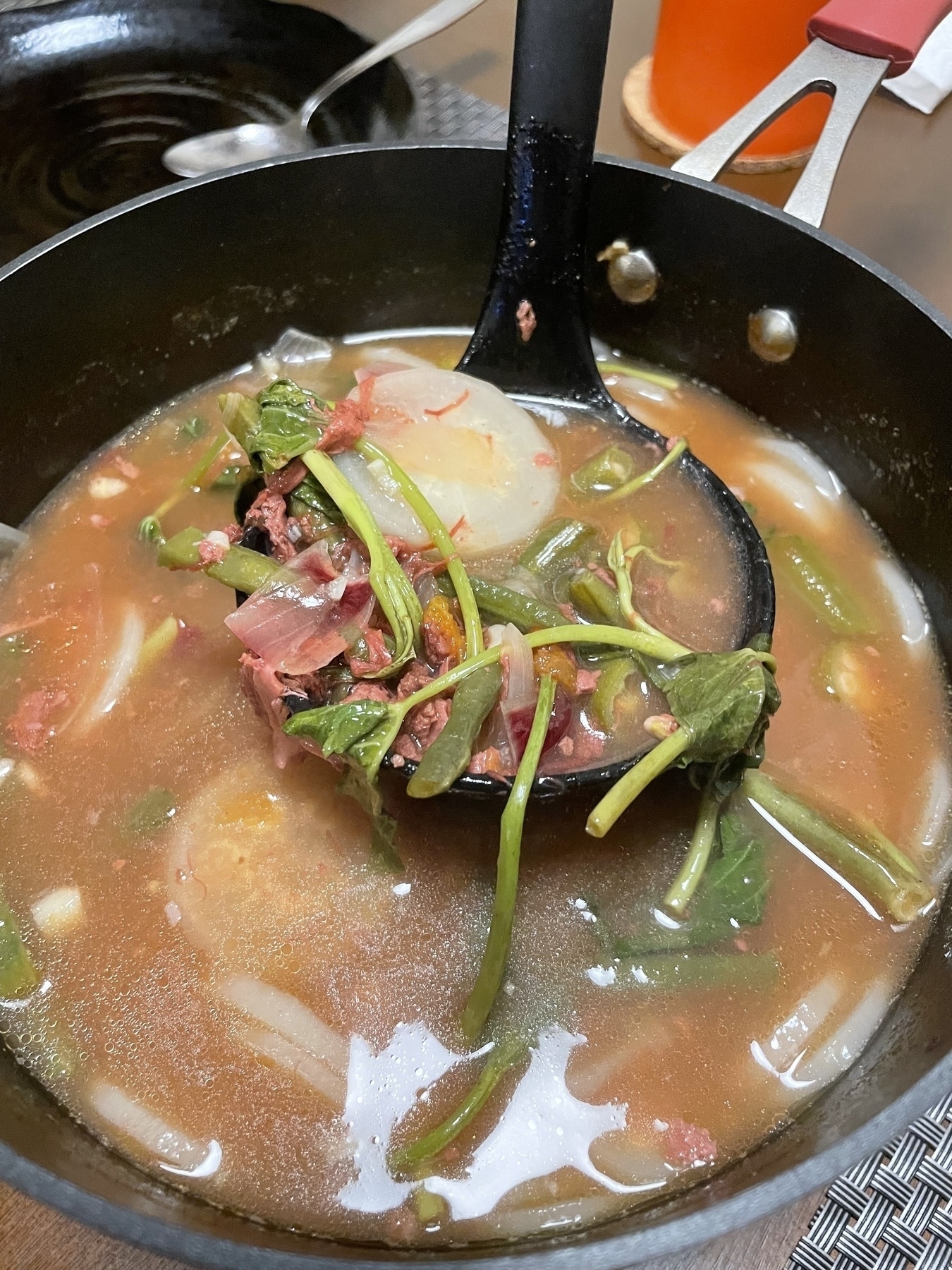 A pot of corned beef sinigang with the ladle holding up a spoonful, containing kangkong, radish, onion bits, and a bit of corned beef.