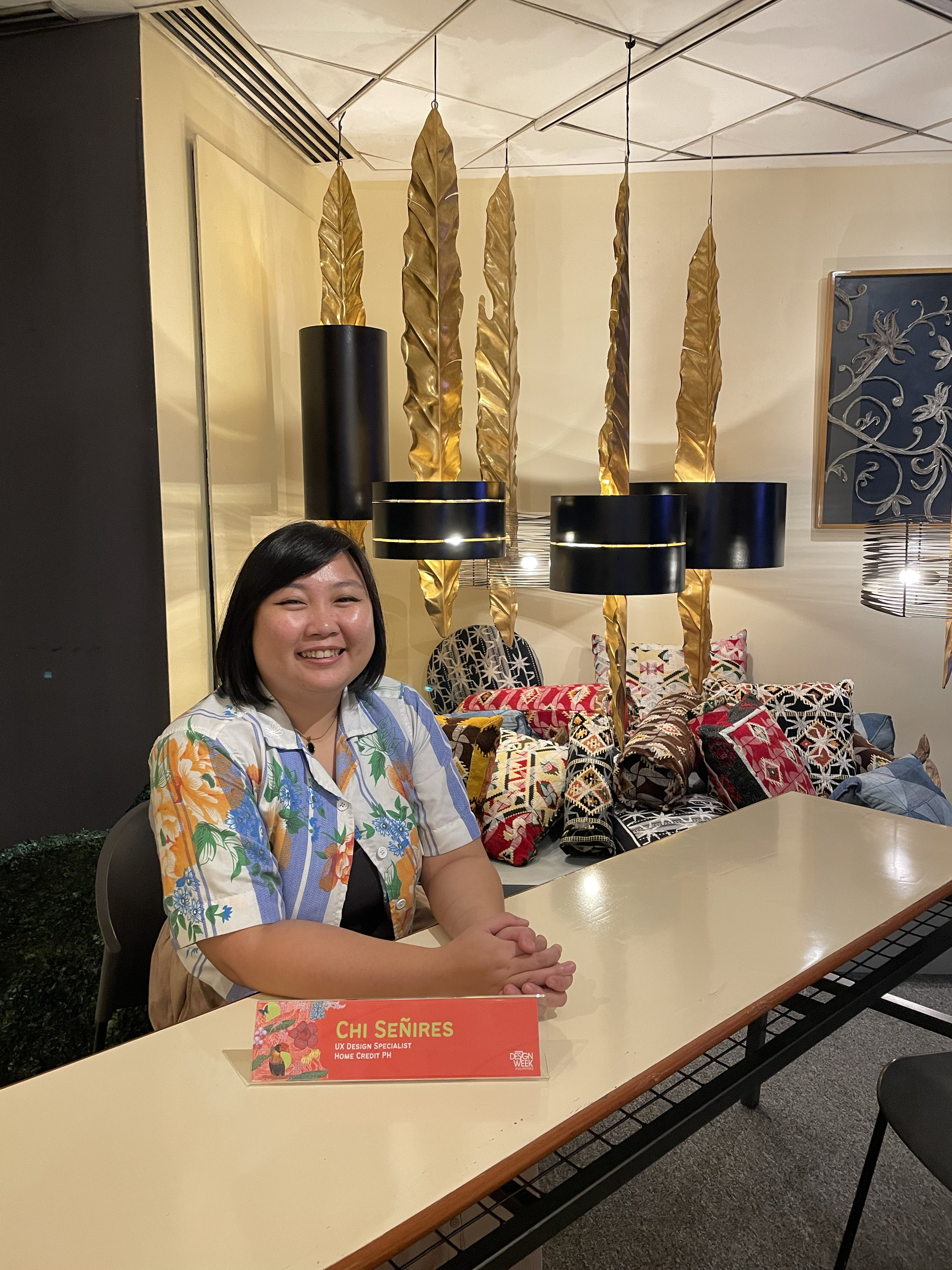 Chi smiling and seated behind a table with her name plate in front of her. Her vibrant floral crop top polo shirt stands out against the elegant decor, including suspended gold leaf decorations and patterned throw pillows.